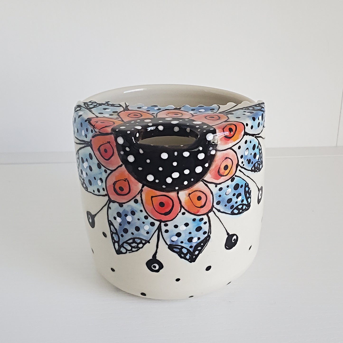 A handmade ceramic planter pot with a white base decorated with a colorful floral mandala pattern in blue, orange, and black, featuring a central hole for a candle holder.