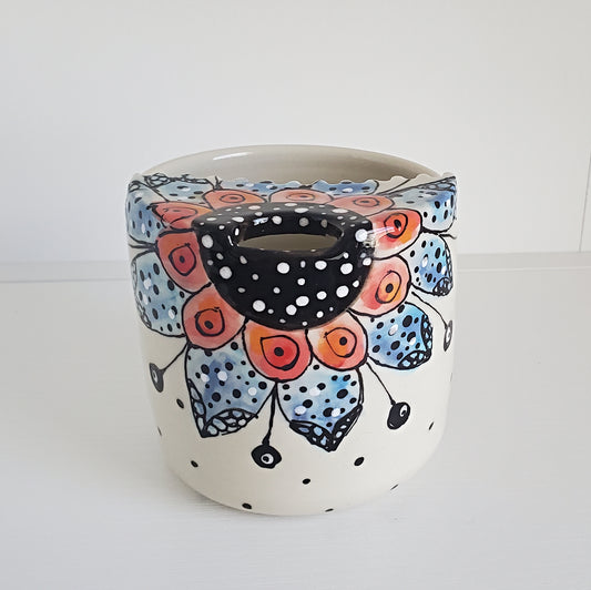 A handmade ceramic planter pot with a white base decorated with a colorful floral mandala pattern in blue, orange, and black, featuring a central hole for a candle holder.