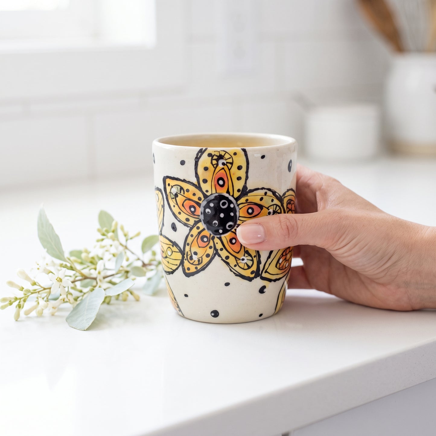 lifestyle: A new photorealistic photograph, composed from scratch, featuring a ceramic cup as the central product. The scene is a clean, bright white minimalist kitchen counter. A small sprig of delicate white and pale green blooms lies next to the product. The overall atmosphere is uplifting, cheerful, and minimal, with cool clean tones and a bright white balance, ensuring no yellow cast. The product is held gently by a woman's hand, positioned slightly off-center to the right, occupying approximately one…